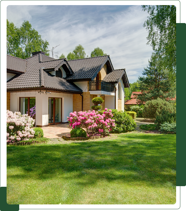 Group 1 (4) A two-story house with a dark roof, surrounded by a lush green lawn and colorful flowering bushes, under a partly cloudy sky.